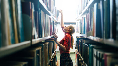 Eine Frau in einem roten Rollkragenpullover und einem karierten Rock steht zwischen zwei Bücherregalen. Sie ist dem linken Bücherregel zugewandt, aus dem sie gerade ein Buch entnimmt.