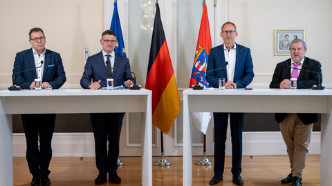 Dr. Christian Höftberger, Ministerpräsident Boris Rhein, Gesundheitsminister Kai Klose und Frank Dastych stehen vor drei Fahnen und halten eine Pressekonferenz ab. Sie stehen an Stehtischen und haben Mikrofone vor sich.