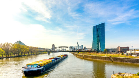 Der Sitz der Europäischen Zentralbank (EZB) im Ostend von Frankfurt am Main, dazu im Vordergrund der Main mit Containerschiff sowie im Hintergrund die Skyline von Frankfurt am Main