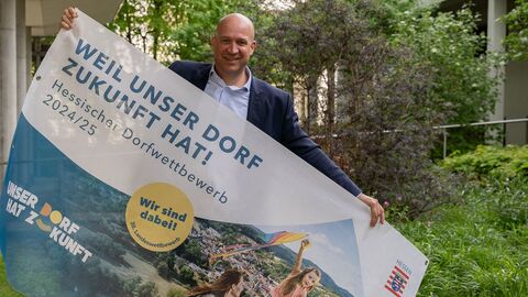 Heimatminister Jung hält ein Plakat für den Dorfwettbewerb in die Höhe, im Hintergrund ist eine Grünanlage zu sehen.