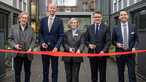 Fünf Personen stehen vor einem Gebäude mit jeweils einer Schere in der Hand. Vor ihnen ein rotes Band welches sie gemeinsam durchschneiden.
