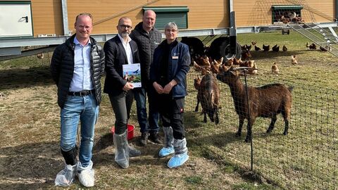 Vier Personen stehen neben einer Weide mit Ziegen und Hühnern, im Hintergrund ist ein Hühnerstall zu sehen.