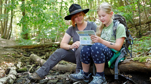 Erwachsene und Kind sitzen im Wald und lesen auf einem Infozettel