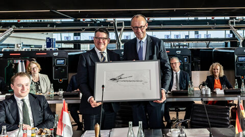 Ministerpräsident Rhein und Bundeskanzler Merz stehen nebeneinander und halten ein gerahmtes Bild.