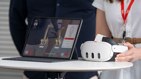 Auf einem Stehtisch steht ein Tablet, dahinter stehen zwei Personen. Eine Person legt eine VR Brille zum Tablet auf den Tisch.