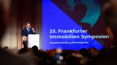 Blick in einen Konferenzsaal: Ministerpräsident Rhein steht vor Publikum an einem Rednerpult auf der Bühne und spricht. Im Hintergrund läuft eine Präsentation mit der AUfschrift "25. Frankfurter Immobilien Symposium".