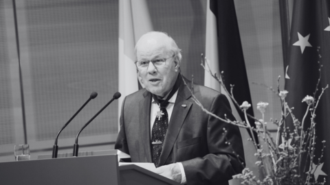 Foto in Schwarz-Weiß: Wolgang Hamberger steht an einem Rednerpult und spricht, hinter ihm sind Flaggen zu erkennen.